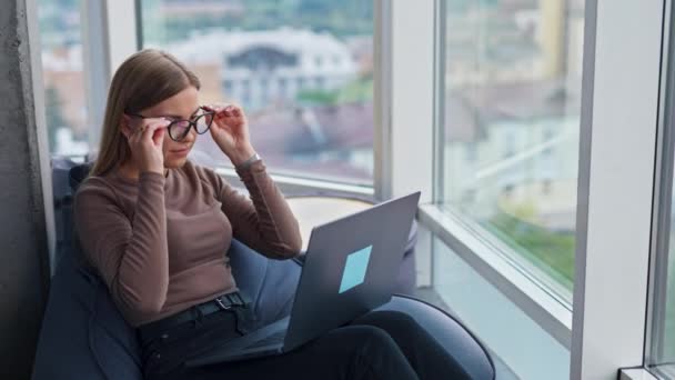 ブロンドの女性は ラップトップで仕事を仕上げメガネを取り外します 疲れた若い女性はコンピュータを閉じ 笑顔の前に窓を見る — ストック動画