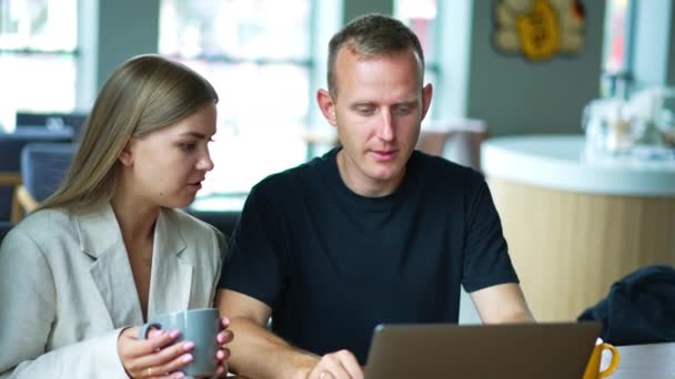 情侣们坐在咖啡馆里手里拿着杯子中年年轻人在笔记本电脑上工作 讨论工作 模糊的背景 — 图库视频影像