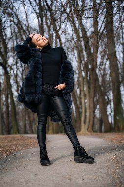 Havalı siyah pantolonları, şık siyah tişörtleri ve şık deri çizmeleri olan kürklü genç bir kadın parkta yürüyor. Havalı kız sonbahar parkında yürüyor..