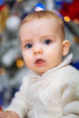 Sevimli erkek bebek Noel ağacının önünde oturuyor ve şaşırmış bir ifadeyle kameraya bakıyor..