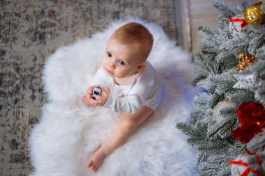 Küçük çocuk Noel ağacının yanındaki beyaz halıda yatıyor ve elinde Noel oyuncağı tutuyor..