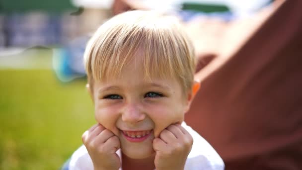 青い目をしている小さな金髪の子供は笑顔で舌を遊ばせます 陽気な天気で屋外のビーンバッグチェアに座っている愛らしい少年 — ストック動画