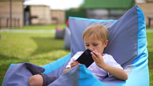 Όμορφο Αγοράκι Αναπαυτική Καρέκλα Κοιτάζοντας Smartphone Στα Χέρια Του Παιδί — Αρχείο Βίντεο