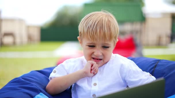 ビーンバッグの椅子の外に座っているかわいい青い目をした少年は 膝の上にラップトップを持っています 子供はスクリーンを見て笑顔 ブラーレッド バックドロップ — ストック動画