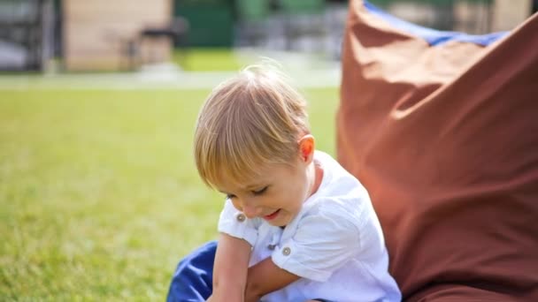 夏に屋外に座っている6歳の美しい髪の男の子 笑顔を見下ろしているシーガール サニーバックドロップ — ストック動画