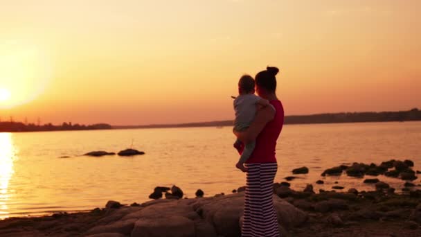 日落时 抱着婴儿的女人站在河边 在大自然中度过了快乐的家庭时光 粉红天空背景 — 图库视频影像