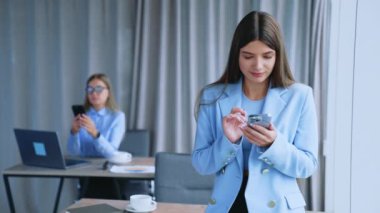 Güzel esmer kadın, elinde telefonla pencerede dikiliyor. Sarışın meslektaşım masanın arkasında oturmuş akıllı telefonuna bakıyor. Bulanık arkaplan.