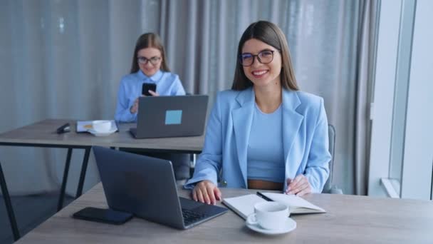 明るい部屋で働く女性社員の幸せ 女性はラップトップとスマートフォンを使用して机に座ります ブラーレッド バックドロップ — ストック動画