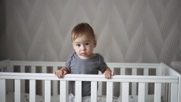 冷静にベビーベッドに立っているカルム コーカサス人の赤ん坊 子供はカメラを覗き込んで 可愛らしい笑顔を浮かべている ライトバックドロップ — ストック動画