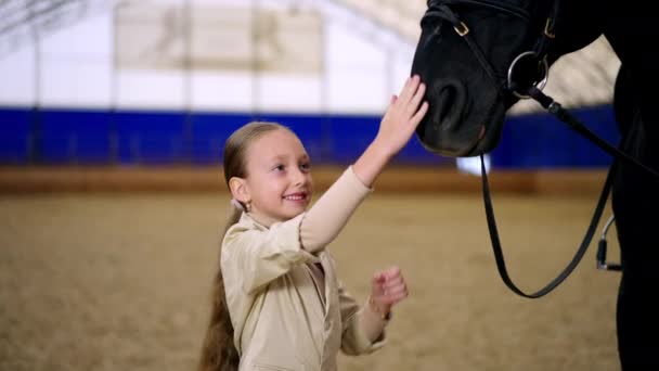 笑笑的白人女孩摸着马的鼻子骑在马鞍上的大黑马走了 在模糊的背景下骑行大厅 — 图库视频影像