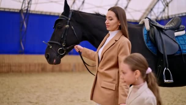 迷人的女士牵着一个女孩的手牵着一匹黑马骑着马鞍和弹药 背景模糊的大型骑车馆 — 图库视频影像