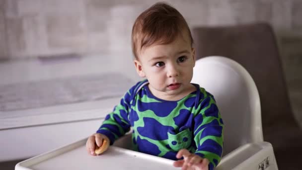 身穿绿色斑点衬衫的活泼可爱的男婴坐在餐桌旁 小男孩挥挥手 然后盯着什么东西看 靠近点 — 图库视频影像