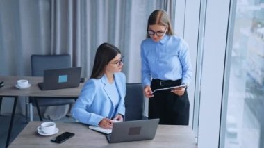 Bayan takım arkadaşları belgelere bakarak iletişim kuruyor. İşyerinde iş birliği yapan, bilgisayar kullanan kadınlar. Yüksek açı görünümü.