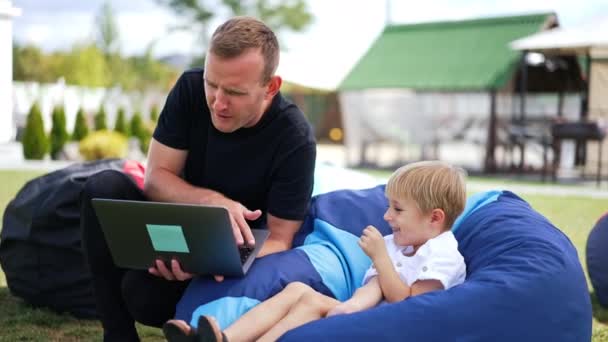 父と息子は屋外で時間を過ごしています ビーン バッグの椅子に座っている少年は 父親の手でラップトップを見ている ブラーレッド バックドロップ — ストック動画