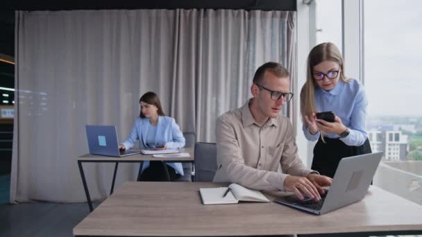 現代のオフィスで働く忙しい人たち 従業員が仕事の問題を解決する コンピュータの前で男性の前に立っている携帯電話を持つ女性 バックドロップのデスクでブルネット女性 — ストック動画