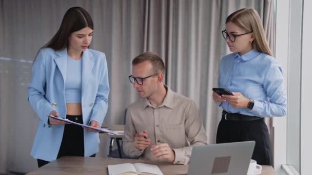 近代的なオフィスでの締め切り時間 デスクに座っている忙しい男性マネージャーと彼の隣に立っている2人の女性従業員 文書に署名する男 — ストック動画