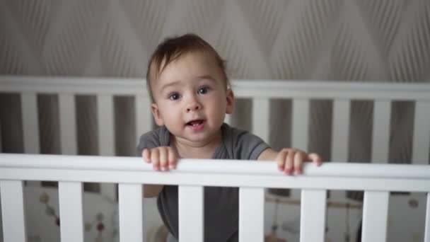漂亮而好奇的小孩站在白色的婴儿床上 可爱的婴儿看着镜头 露出舌头 — 图库视频影像