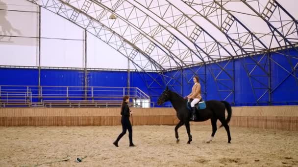 美しい黒い馬に乗っている長髪の女性 女性のコーチが動物の前を歩いています 若い女の子が前景に上がってくる — ストック動画