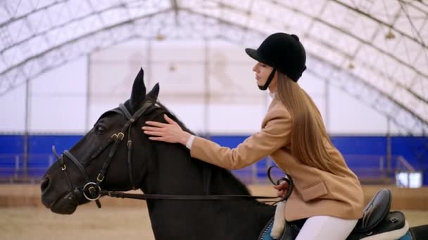 身穿黑色头盔和米黄色夹克的女骑师骑着一匹漂亮的黑马 女人让爱抚它的动物平静下来 模糊的背景 — 图库视频影像
