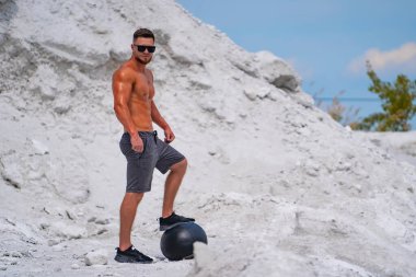 Kaslı adam beyaz kumsalda fitness topuyla poz veriyor..