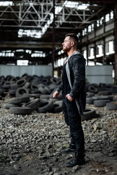 Man stands in black jacket and jeans in abandoned building with old tires.