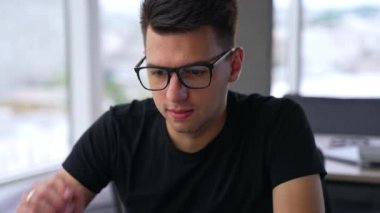 Koyu renk saçlı yakışıklı adam bilgisayar ekranına daha yakından bakmak için gözlüklerini çıkarıyor. Büyük ışık odasında dizüstü bilgisayarda çalışan serbest çalışan biri. Bulanık arkaplan.