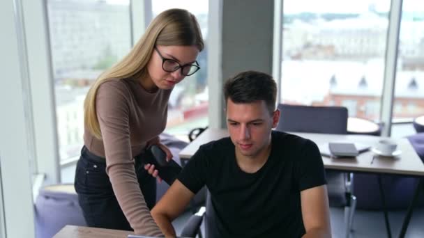 メガネをかけている女性は ノートパソコンで働く男性の近くに立っています 女性はそれをコメントする画面を指します バックドロップでぼやけたパノラマウィンドウ — ストック動画