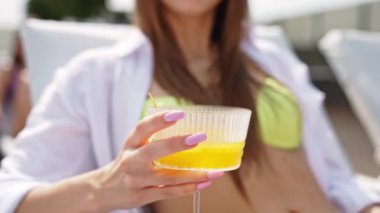 Sarı kokteylden hoşlanan çekici bayan. Beyaz tenli genç bir kadın yaz mevsiminde bir arkadaşıyla güvertede güneşleniyor..