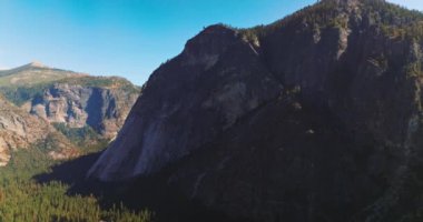 Tepelerinde ve ayaklarında çam ağaçları olan büyük dağlar. Yosemite Ulusal Parkı açık mavi gökyüzünün zemininde sallanıyor.