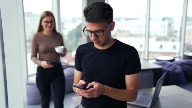 Öğle yemeği molasında iş arkadaşları ofiste. Elinde telefon olan bir adam ve ona yardım etmek için bir kadın geliyor. Görüntü arkaplanındaki panoramik pencereler.