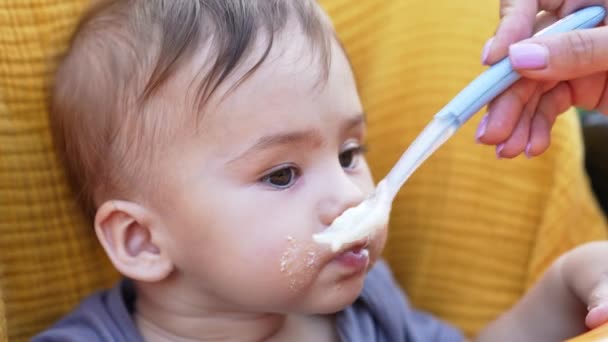 スプーンから食べる頬に食べ物を持った素敵な白人子供 母の手は赤ちゃんにお粥をあげている クローズアップ — ストック動画