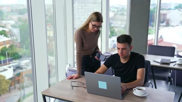 オフィスで働く二人が協力している 男はラップトップに座り 女はスクリーンを指さすトークの横に立っている ブラーレッド バックドロップ — ストック動画