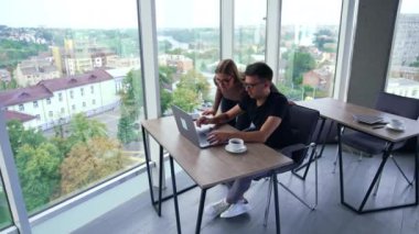 Bir çift meslektaşımız dikkatle dizüstü bilgisayara bakıyor. Meslektaşlar bilgisayarda bazı işlerle meşguller. Pencerelerinde şehir manzarası olan kapalı bir ofis arkaplanı.