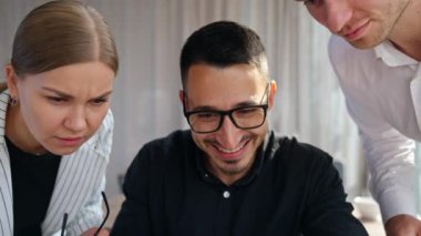Gözlüklü mutlu bir erkek dizüstü bilgisayarın önünde oturuyor. Yanında duran iki meslektaşı dikkatle ekrana bakıyor ve konuşuyor..