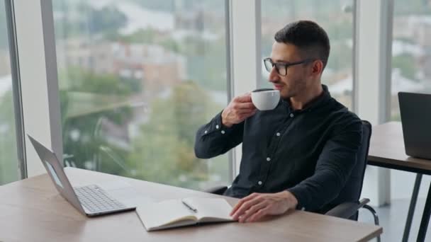 ブラックシャツのブルネット男が 仕事場でコーヒーを飲んでる 窓を見る仕事から休憩している男性従業員 — ストック動画