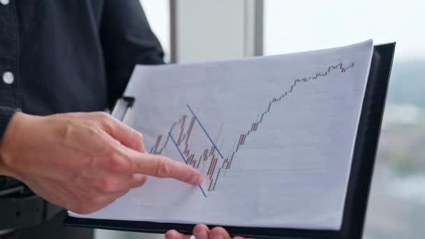 紙に印刷されたグラフに従う男性の手 黒服のビジネスマンがチャートを指して話す オフィスの屋内背景 — ストック動画