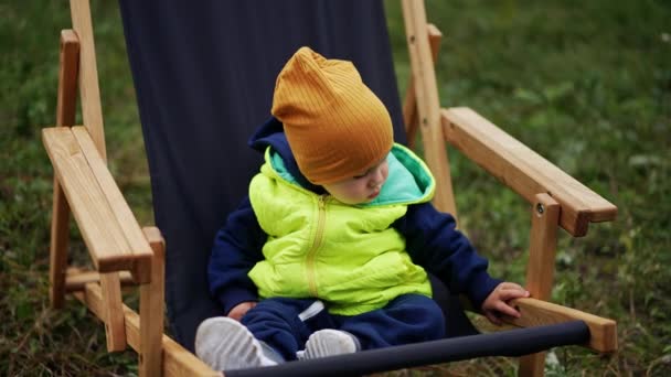 白种人小孩穿着暖和的衣服 戴着一顶帽子 坐在花园的椅子上 婴儿小心地四处张望 安详地坐着 靠近点模糊的自然背景 — 图库视频影像