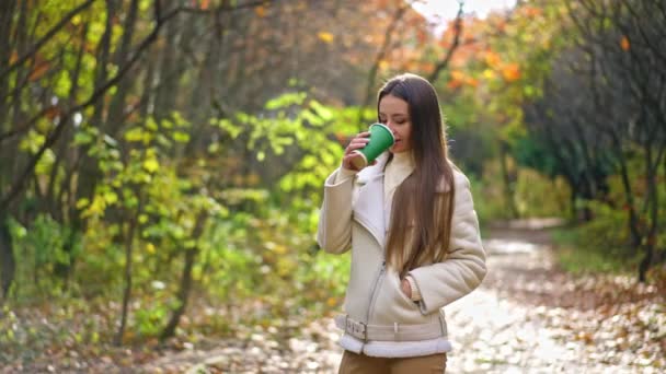 身穿乳白色夹克的黑头发女士在公园散步时喝咖啡 秋天里一个人散步的女人 — 图库视频影像