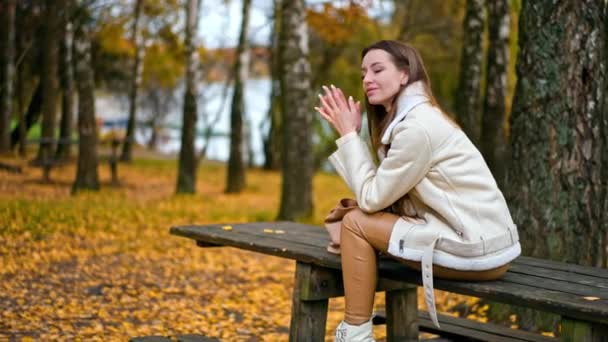 落ち着いた落ち着いた暗い髪の女性は 何かを考えているテーブルの上に座ります 鳥の木の間で公園で美しい秋の日を楽しんでいる白人少女 — ストック動画