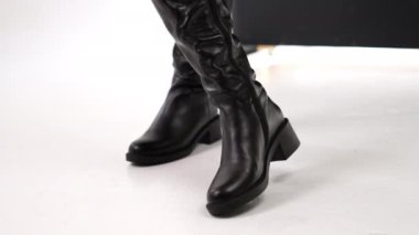 Düz tabanlı ve alçak topuklu parlak siyah yüksek deri çizmeler. Ayakkabıları göstermek için beyaz zeminde dönen bir model. Kapat..