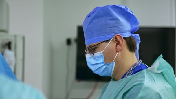 中年男性外科医生身穿蓝色制服 头戴外科口罩 头戴帽子和眼镜 站在旁边看着前方的摄像机 在手术室工作的医生小组 — 图库视频影像