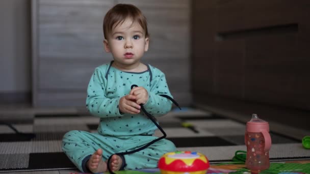 黒いレースを手に持っている床に座っている青いパジャマの小さな子供 自宅で床で遊んでいる素敵な子供 ブラーレッド バックドロップ — ストック動画