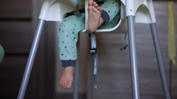 高い椅子に座っている赤ちゃんの愛らしい小さな裸足 青いズボンの子供が足を振っている クローズアップ ブラーレッド バックドロップ — ストック動画