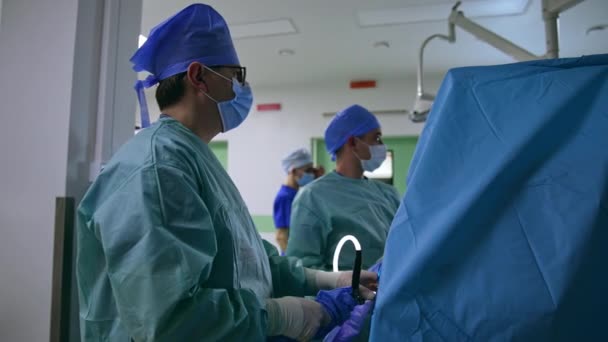 メガネの大人の男性医師は 2つのチューブで操作を行っている器械を保持し 動かします 外科医とアシスタントがスクリーンを見る — ストック動画