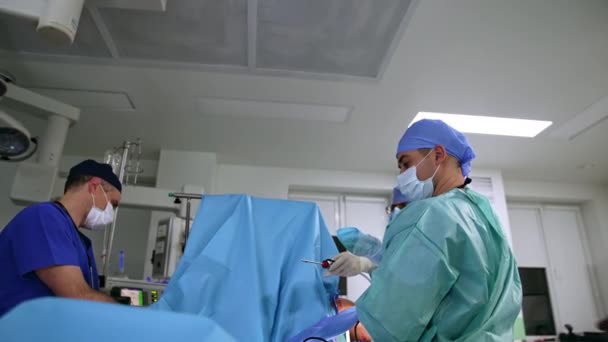 若い男性アシスタントは 外科医からツールを取って デバイスをチーフドクターに渡します 現代の診療所で患者を操作する医療専門家のチーム ローアングルビュー — ストック動画