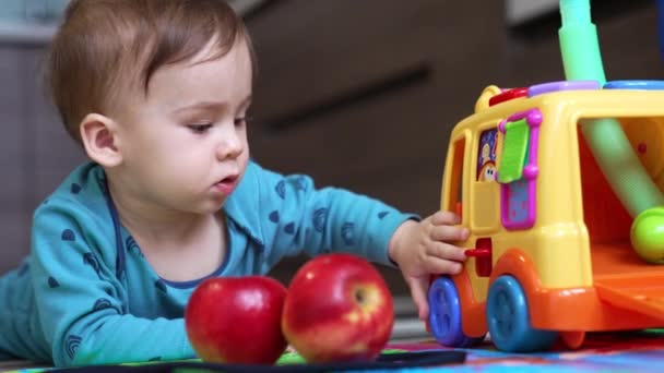 甘い素敵な子供は 彼の前にリンゴを持って床に横たわっています 明るいおもちゃの車に触れ 車のドアを開け 閉めるのに適した幼児 クローズアップ — ストック動画