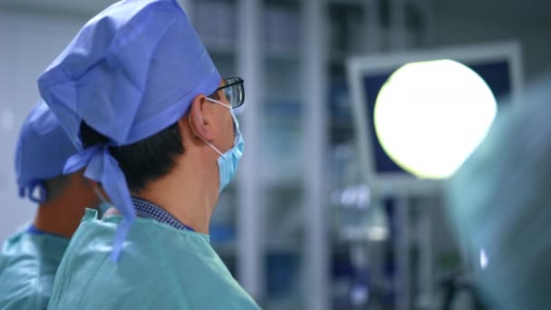 目の前でモニターを眺めている眼鏡をかぶった注意深い外科医 スクリーンに焦点を当てた医師のリアビュー ブラーレッド バックドロップ — ストック動画