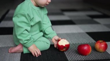 Açık yeşil elbiseli en tatlı beyaz çocuk dizlerinin üzerine çöker. Bebek, yere saçılmış elmalardan ısırılmış elma alır. Bulanık arkaplan.