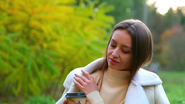 暖かい秋の自然のバイブを楽しむ 見栄えの良い白人女性 ブルネットは彼女の手に紙コップで地面に座る — ストック動画