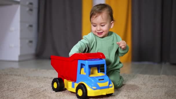 彼の前の床に明るいおもちゃのローリーを持つ幸せな愛らしい子供 赤ん坊の少年は車のバスケットから赤いボールを取り出し 元気に投げる — ストック動画